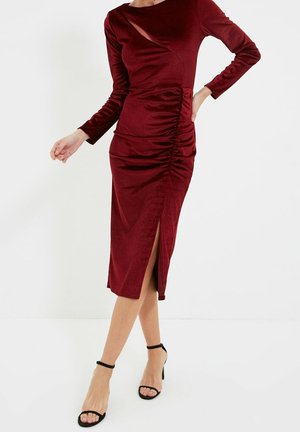 Femme portant une robe midi moulante en velours bordeaux à manches longues avec une fente latérale et un détail froncé, associée à des talons noirs à bride à la cheville.