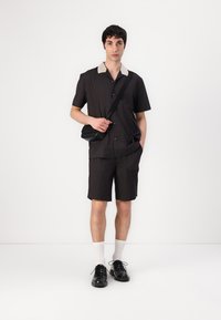 Chemise noire à rayures avec des manches courtes et un col de couleur claire, shorts assortis, chaussures noires et un sac en bandoulière. Chaussettes blanches visibles.