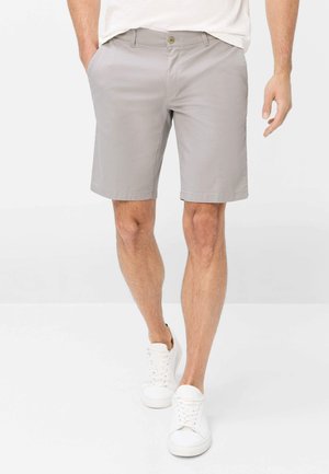 Mand iført lysegrå knælange shorts, hvide sneakers og en hvid skjorte, stående med hænderne i lommerne på ensfarvet baggrund.