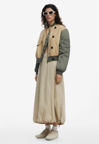 Beige and green bomber jacket, oversized, with buttons and zip closure. Flowing beige skirt, ankle-length, with gathered hem. Light-colored shoes.