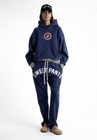 Sweat-shirt à capuche bleu marine avec un logo circulaire, associé à un pantalon de jogging assorti avec l'inscription en lettres audacieuses "SWEET PANTS". Tissu doux, coupe décontractée.