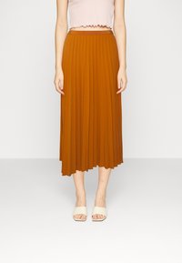 Jupe midi plissée marron en tissu léger, dotée d'une taille haute et d'une texture lisse. Associée à des chaussures blanches ouvertes à l'avant.