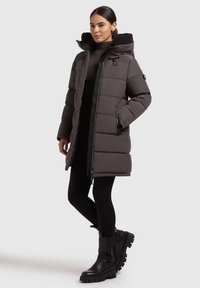 Abrigo gris acolchado con un alto cuello y capucha removible, cierre de cremallera, bolsillos laterales y tejido texturizado. Combinado con leggins negros y botas.