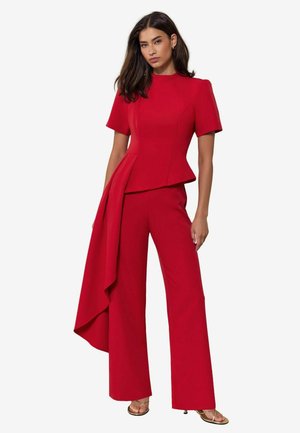 Woman wearing a red short-sleeve peplum top and matching wide-leg pants with a draped panel on one side, paired with gold open-toe heels.