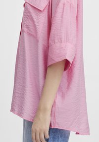 Camicetta corta rosa con finitura testurizzata, caratterizzata da maniche corte e una vestibilità ampia. Le spaccature laterali migliorano i movimenti; il tessuto sembra leggero.