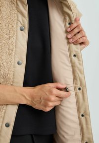 Beige gesteppte Jacke mit einer strukturierten, kunstpelzähnlichen Außenseite. Schwarze Druckknöpfe und eine verstellbare Kordelzugdetails im Innenfutter.