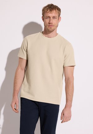 Homme aux cheveux blonds courts, portant une chemise beige à manches courtes et un pantalon bleu marine, debout devant un fond clair uni.
