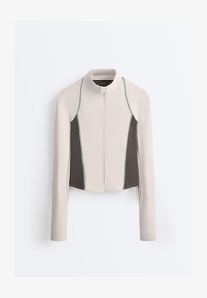 Langærmet beige jakke med lynlås, mørkegrå sidepaneler og ståkrave, vist mod en hvid baggrund.