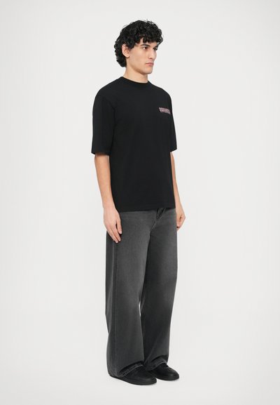 Črna bombažna majica s kratkimi rokavi in okrolim izrezom, z majhnim roza logotipom na prsih. Kombinirana z ohlapnimi sivimi jeans hlačami.