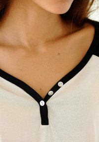 White top with black accents, features a v-neck, ribbed collar, and three silver buttons at the neckline, showcasing a smooth texture.