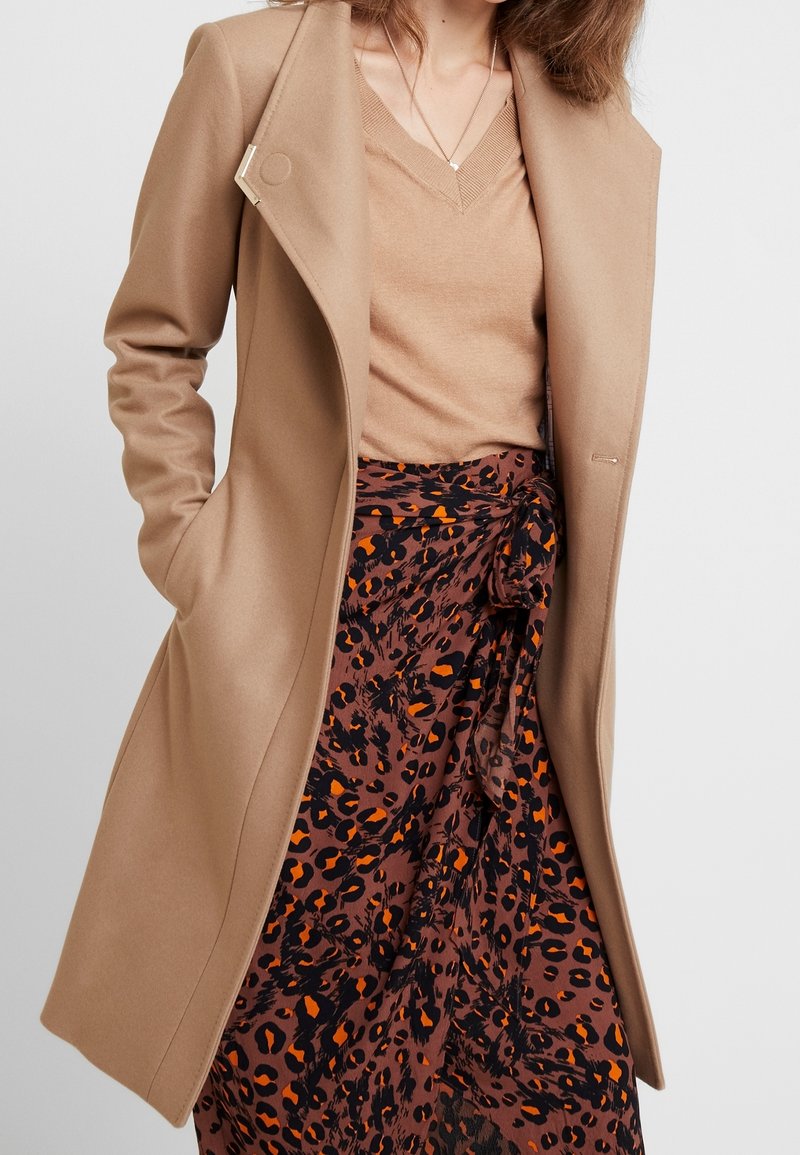 Femme portant un manteau couleur chameau sur un pull beige à col en V et une jupe marron avec un imprimé léopard noir et orange, les mains dans les poches du manteau.