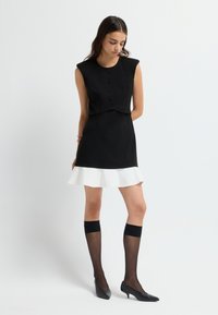 Frau in schwarzem ärmellosem Oberteil mit Knöpfen, schwarzem Minirock mit weißem Rüschensaum, schwarzen transparenten knielangen Socken und schwarzen High Heels.