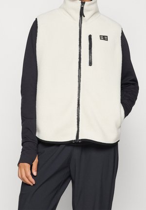 Henkilö pukeutunut valkoiseen fleeceliiviin, jossa on musta vetoketju ja rintatasku, mustan pitkähihainen paidan päällä, seisoo yksiväristä taustaa vasten.