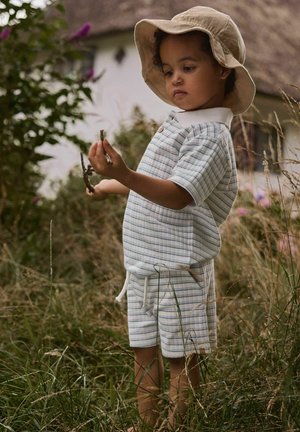 Bambino piccolo con cappello da sole e abito a righe sta in piedi nell’erba alta, tenendo e esaminando piccoli rami all’aperto vicino a una casa sfocata.