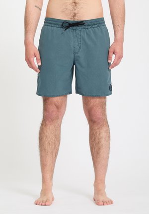 Hombre con pantalones cortos de baño color verde azulado con cordón negro, de pie descalzo contra un fondo blanco liso.