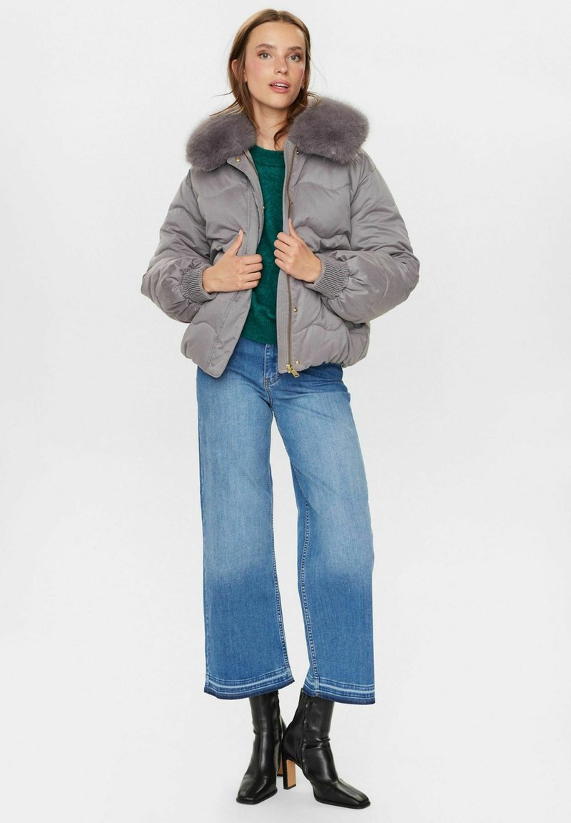 Grau gepolsterte Bomberjacke mit einem Kragen aus Kunstfell, getragen über einem grünen Pullover, kombiniert mit weiten blauen Jeans und schwarzen Knöchelstiefeln.