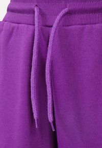 Fialové tepláky mají žebrovaný pas s odpovídajícími stahovacími šňůrkami, texturovanou tkaninu a hladký povrch. Konečky stahovacích šňůrek jsou plastové.