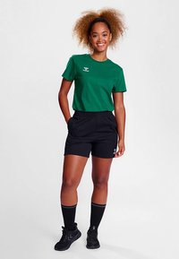 Camiseta verde de manga corta con logo, combinada con pantalones cortos negros con bolsillos laterales. Medias negras hasta la rodilla y zapatos deportivos negros completan el conjunto.