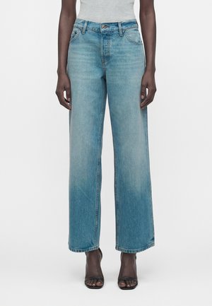 Kvinde iført høj talje, lyseblå jeans med lige ben kombineret med sorte sandaler med åben tå og en grå ærmeløs top.