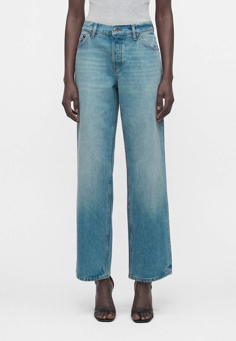 Γυναίκα φοράει τζιν ψηλόμεσο, ανοιχτού μπλε χρώματος με ίσιο μπατζάκι, συνδυασμένο με μαύρα πέδιλα με ανοιχτά δάχτυλα και γκρι αμάνικη μπλούζα.