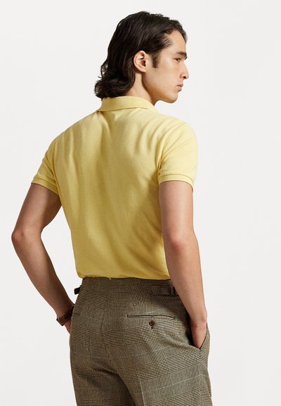 Dzeltena polo krekla ar īsām piedurknēm, ar teksturētu audumu. Redzamas brūnas svītrotas bikses ar aizmugurējām kabatām un jostas cilpiņām.