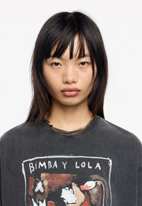 Camiseta gris de gran tamaño con un diseño gráfico colorido que incluye elementos florales. La camiseta se combina con un delicado collar de cuentas multicolores.