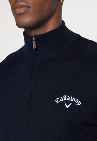 Pull-over en maille bleu marine avec col zippé. Comprend un logo "Callaway" blanc brodé sur la poitrine gauche et une texture côtelée.