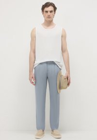 Uomo che indossa una maglietta senza maniche bianca, pantaloni azzurri chiari, scarpe beige senza lacci, e tiene un cappello di paglia con strisce blu nella mano destra.