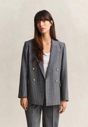 Frau mit langen braunen Haaren, die einen grauen Nadelstreifen-Doppelreiher-Blazer mit goldenen Knöpfen und passende Hose trägt, stehend vor neutralem Hintergrund.