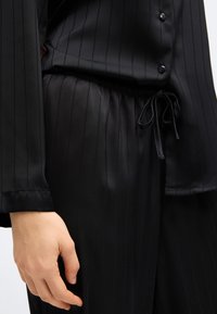 Κοντινό πλάνο ενός ατόμου που φοράει μαύρο παντελόνι σατέν με ρίγες, ελαστική μέση και κορδόνι, συνδυασμένο με ένα μαύρο πουκάμισο με κουμπιά που ται ριάζει.