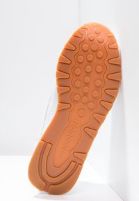 Witte sneaker met een leren bovenkant met textuur en een oranje rubberen zool. De zool heeft cirkel- en golfpatronen voor grip. Reebok-logo zichtbaar.