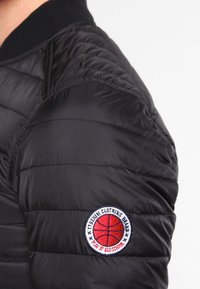 Musta toppatakki, jossa on tikattu kuvio. Ominaisuuksina pyöreä merkki, jossa punainen koripallo ja teksti "PLAY IT OLD SCHOOL."