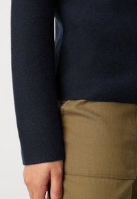 Bras dans un pull-texturé bleu marine à manches longues reposant à côté de la taille, portant un pantalon marron clair avec des détails de couture visibles.