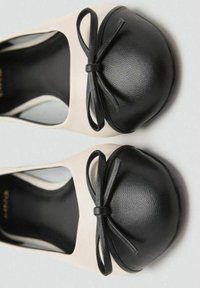 Un par de zapatos planos beige con puntas redondeadas en negro y acentos de lazo negro, mostrados desde arriba sobre un fondo blanco.