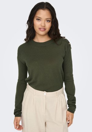 Mujer con suéter de manga larga verde oscuro con detalles de botones en el hombro y pantalones plisados beige de talle alto, de pie frente a un fondo blanco.