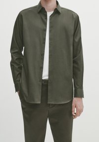 Camicia verde oliva con bottoni, dal tessuto liscio, maniche lunghe e orlo dritto; abbinata a pantaloni scuri coordinati e una maglietta bianca.