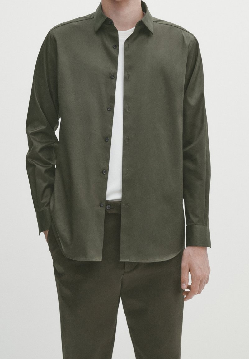 Camicia verde oliva con bottoni, dal tessuto liscio, maniche lunghe e orlo dritto; abbinata a pantaloni scuri coordinati e una maglietta bianca.