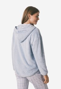 Hellblaue Fleece-Hoodie mit einer weichen, flauschigen Textur, ausgestattet mit einer Kapuze und einem lässigen Schnitt. Kombiniert mit karierten Schlafanzughosen.