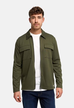 Młody mężczyzna zwrócony przodem, ubrany w otwartą oliwkowozieloną kurtkę na zamek, na białej koszuli i ciemnoniebieskich dżinsach, na jednolitym tle.