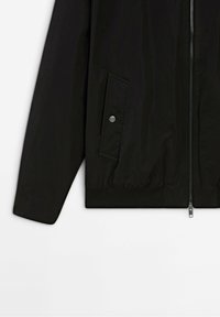 Manche de veste noire avec fermeture éclair à l'avant et une poche à rabat fermée par un bouton-pression argenté sur fond blanc.