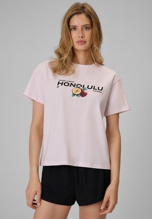 Młoda kobieta z falującymi blond włosami, ubraną w jasnoróżowy t-shirt z napisem Honolulu Hawaii oraz czarne szorty na jednolitym szarym tle.
