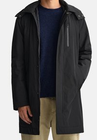 Sort, vandtæt lang frakke med foret foer, lynlåslomme på brystet og krave med trykknapper, båret over en marineblå sweater.