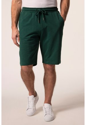 Mand iført mørkegrønne shorts med snøre, med hænderne i lommerne og hvide sneakers mod en ensfarvet beige baggrund.