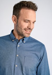 Camisa azul abotonada hecha de una tela texturizada que presenta un bolsillo frontal, cuello abotonado y botones de color claro.