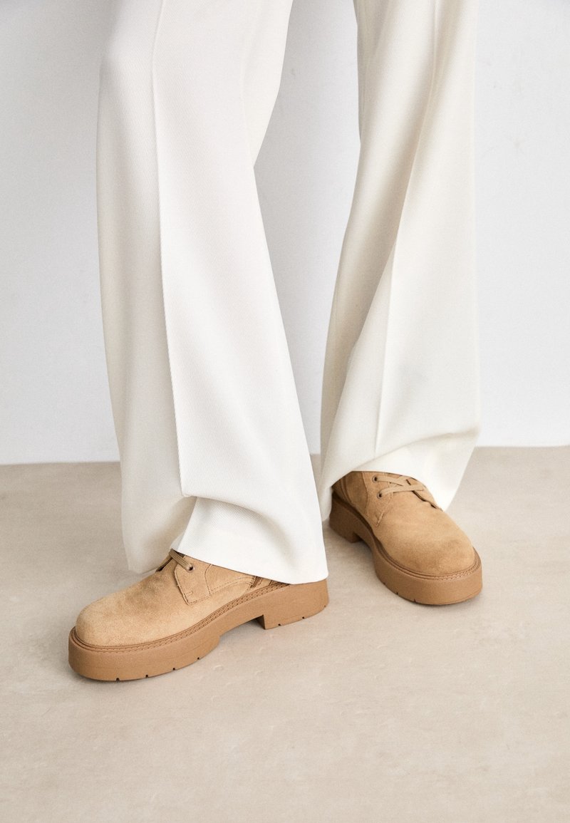 Chaussures en daim beige à lacets portées avec un pantalon large crème sur un sol neutre.