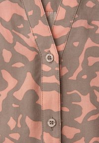 Chemise à manches courtes en tissu marron clair avec un motif camouflage rose. Elle présente un col en V et deux boutons à l'avant.