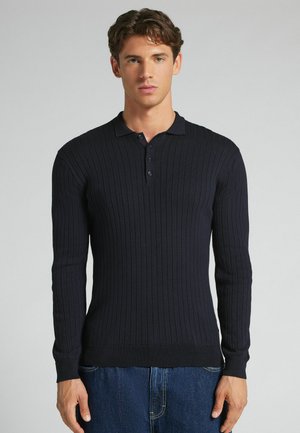 Mann trägt ein schwarzes, geripptes Langarm-Poloshirt und blaue Jeans und steht vor einem schlichten hellen Hintergrund.