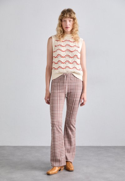 Pull sans manches en tricot avec un motif de vagues en crème et rouge, associé à un pantalon évasé à carreaux marron et des chaussures à lacets beiges.