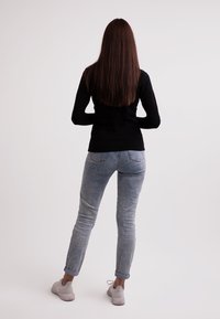Schwarzes Langarm-Oberteil, hellblaue Jeans, hochgerollte Ärmel und helle Sneakers. Das Modell steht mit dem Rücken zur Kamera vor einem schlichten Hintergrund.
