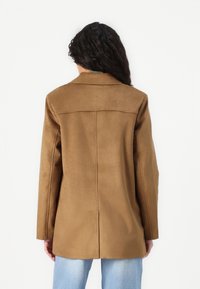 Femme aux longs cheveux bouclés portant une veste en daim marron et un jean bleu clair, vue de dos sur un fond blanc uni.